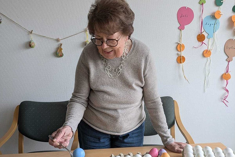 Person steht an einem Tisch mit mehreren Gläsern gefärbter Flüssigkeit, Eierkartons mit gefärbten Eiern und bunten Buchstaben an der Wand.