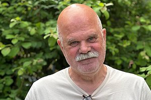 Mann mit weißem T-Shirt und Brille am Hals vor grünem Laub im Freien