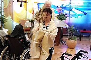 Person in weißem Gewand trägt liturgisches Tuch in einem Raum mit Rollstühlen und buntem Hintergrundfenster