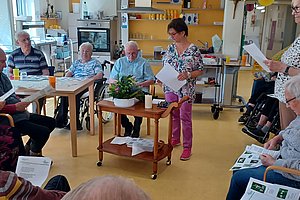 Mehrere ältere Menschen sitzen in einem Raum an Tischen und halten Blätter, zwei Frauen stehen und lesen vor