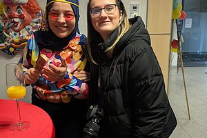 Zwei Frauen in bunten Kostümen lächeln gemeinsam, eine mit Zirkus-Elementen, die andere mit Kamera.