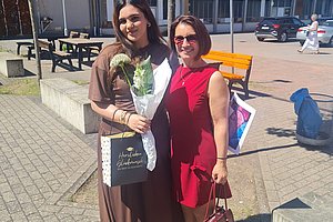 Zwei Frauen stehen im Freien vor einem Gebäude, eine trägt ein langes braunes Kleid und hält einen Blumenstrauß und eine Geschenktüte, die andere trägt ein rotes Kleid und eine Handtasche