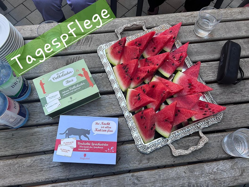 Auf einem Holztisch steht eine silberne Platte mit dreieckigen Wassermelonenstücken, daneben liegen zwei bunte Kartons und mehrere Gläser mit Wasser.