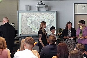 Mehrere Personen stehen vor einem Beamerbild mit Blumenmuster in einem Raum mit Publikum im Vordergrund