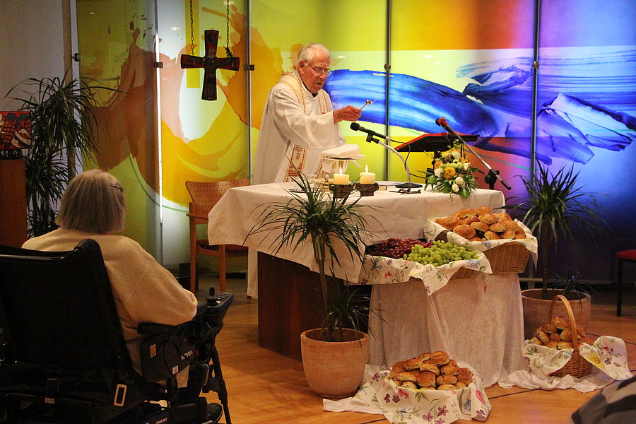 Ein Priester steht an einem mit Brot, Trauben und Kerzen gedeckten Altar, während eine Person im Rollstuhl davor sitzt.