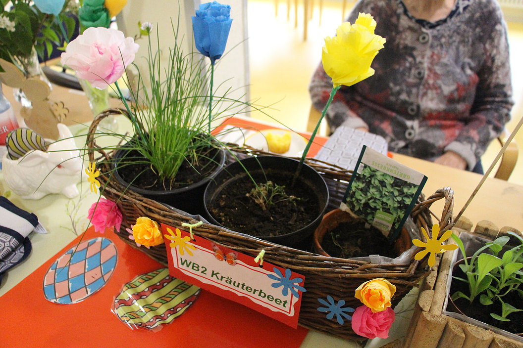 Korb mit drei Kräutertöpfen, dekoriert mit Papierblumen und Schild 'WB2 Kräuterbeet', auf orangem Tisch, im Hintergrund sitzende Person.