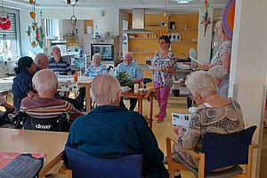 Gruppe älterer Menschen sitzt in einem hellen Raum an Tischen, eine Frau steht und spricht zu ihnen