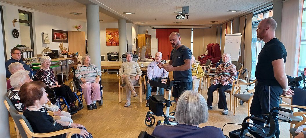 Mehrere ältere Menschen sitzen in einem Stuhlkreis in einem hellen Raum, zwei Männer stehen in der Mitte und sprechen zu der Gruppe.