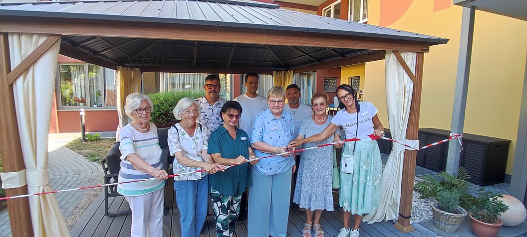 Gruppe älterer Menschen steht unter einem Holzdach und hält gemeinsam ein langes rotes Band