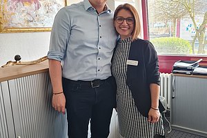 Zwei Personen stehen lächelnd in einem Raum mit Fenstern und einem Schrank im Hintergrund.