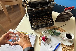 Hände formen ein Herz über einem aufgeschlagenen Buch mit Foto und Text neben einer alten Schreibmaschine, einem Bügeleisen, einer Rose, einem Stift und einer Tasse Kaffee auf einem Tisch