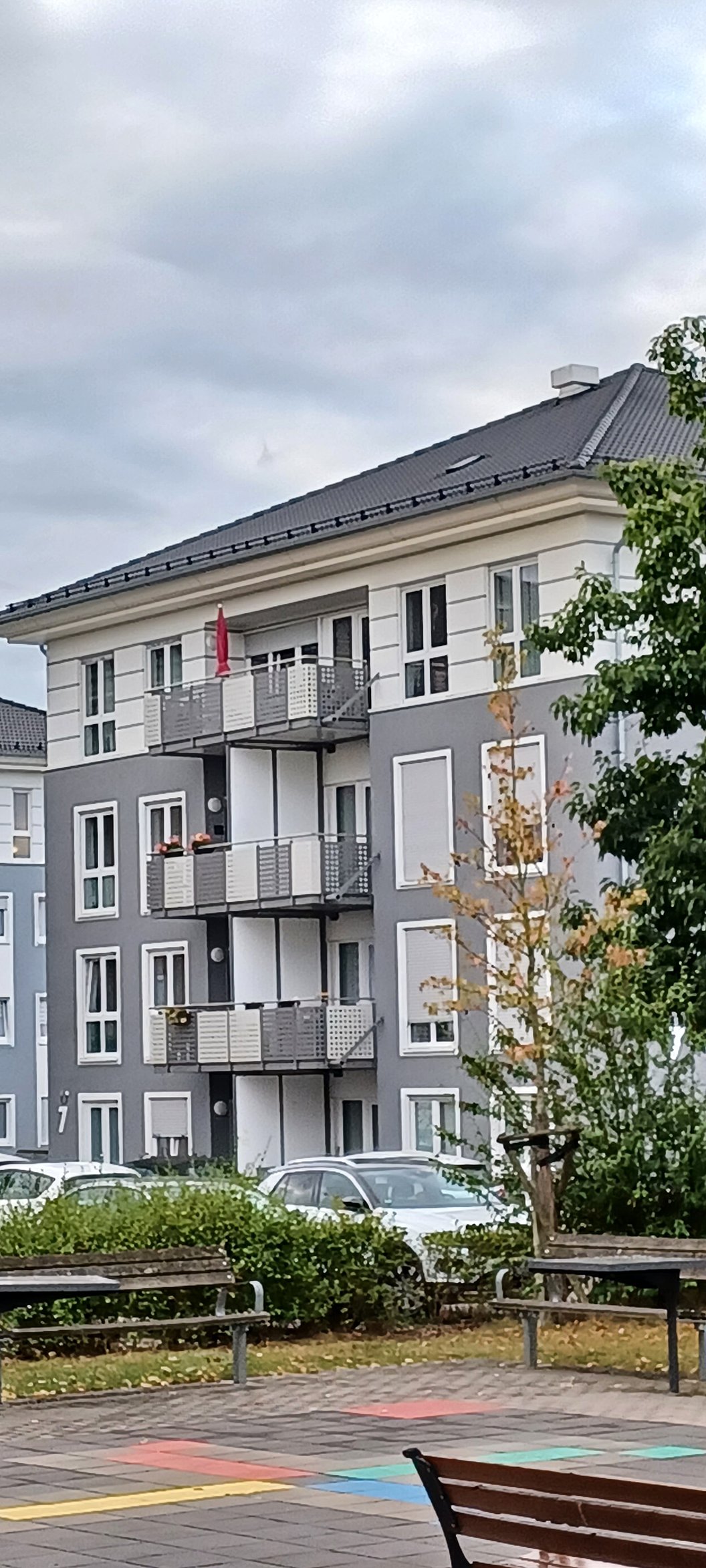 Mehrstöckiges Wohngebäude mit Balkonen, davor Parkbänke und ein kleiner Spielplatz mit buntem Bodenbelag.