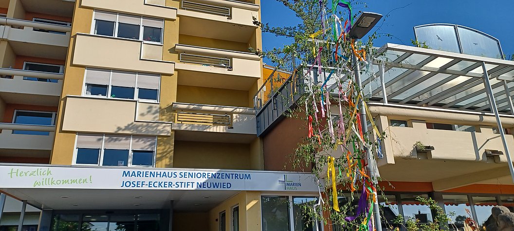 Mehrstöckiges Seniorenwohnheim mit Balkon und Eingangsbereich, davor ein geschmückter Maibaum mit bunten Bändern.