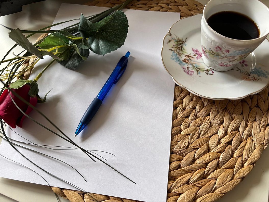 Auf einem geflochtenen Untersetzer liegt eine Tasse Kaffee auf einer Untertasse mit floralem Muster neben einem blauen Kugelschreiber und einer roten Rose auf weißem Papier