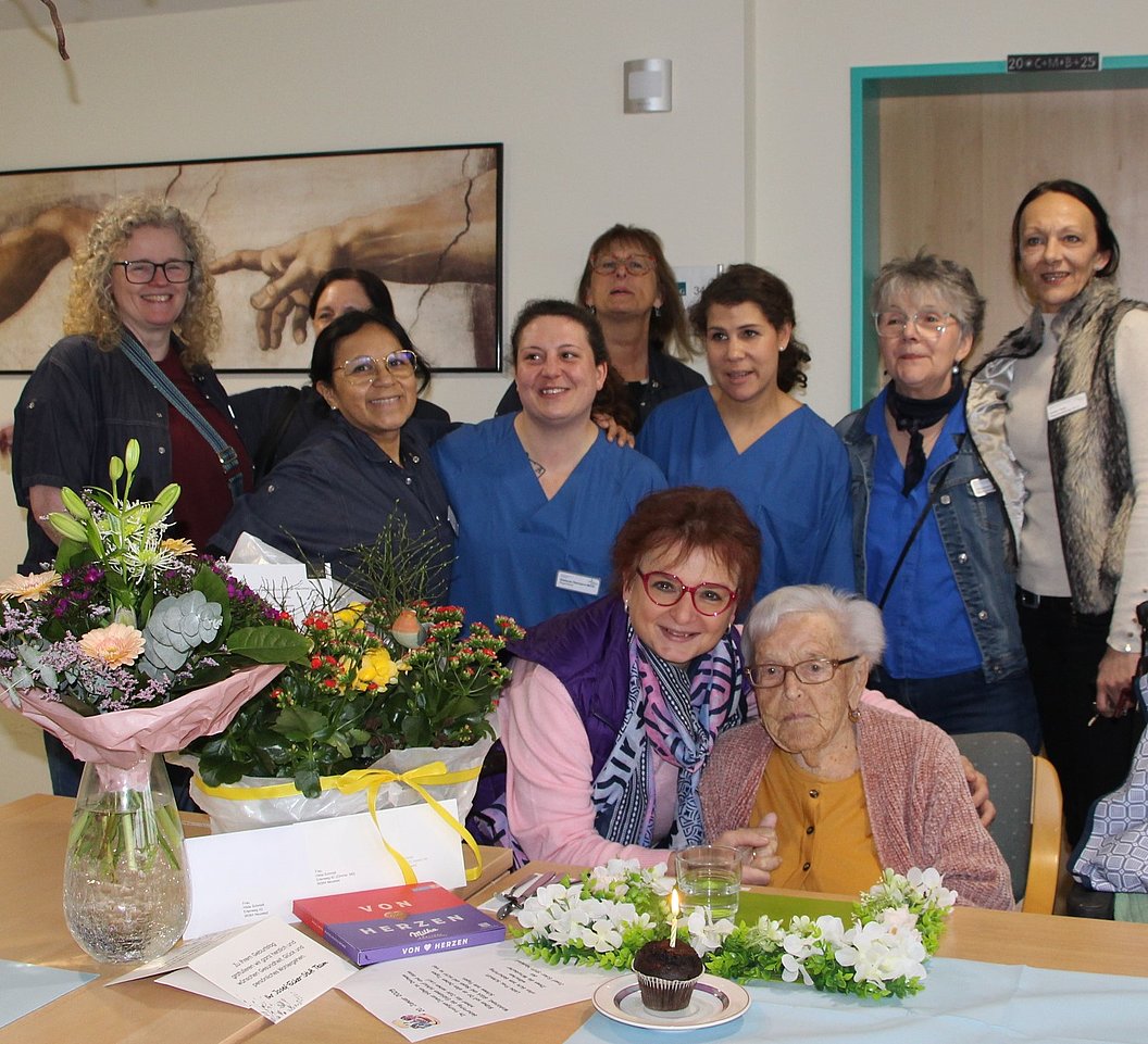 Gruppe von Menschen in einem Raum mit einem Tisch, auf dem Blumensträuße, Karten und ein kleiner Kuchen mit Kerze stehen.