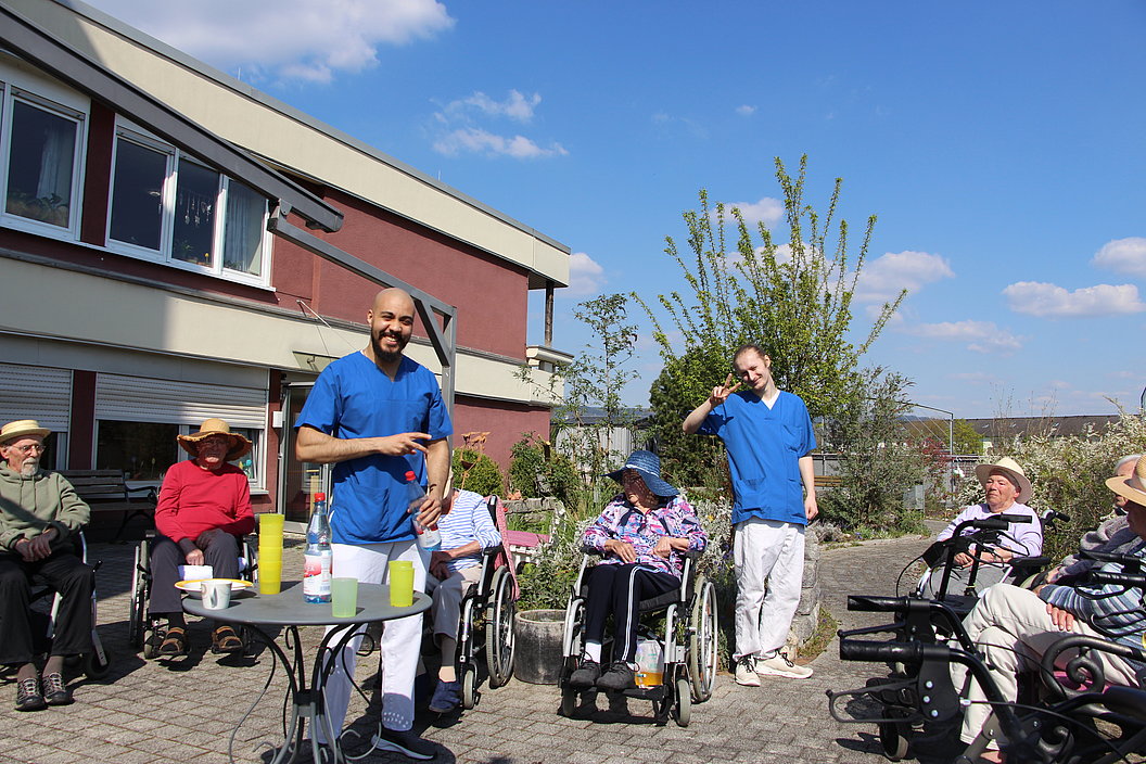 Mehrere ältere Menschen, einige im Rollstuhl, sitzen im Außenbereich eines Gebäudes, während zwei Personen in blauer Arbeitskleidung mit ihnen interagieren.