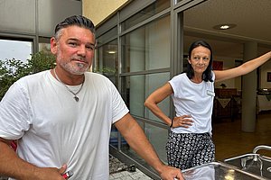 Zwei Personen in weißem T-Shirt stehen an einem Edelstahlwagen vor einem Gebäude mit großen Fenstern