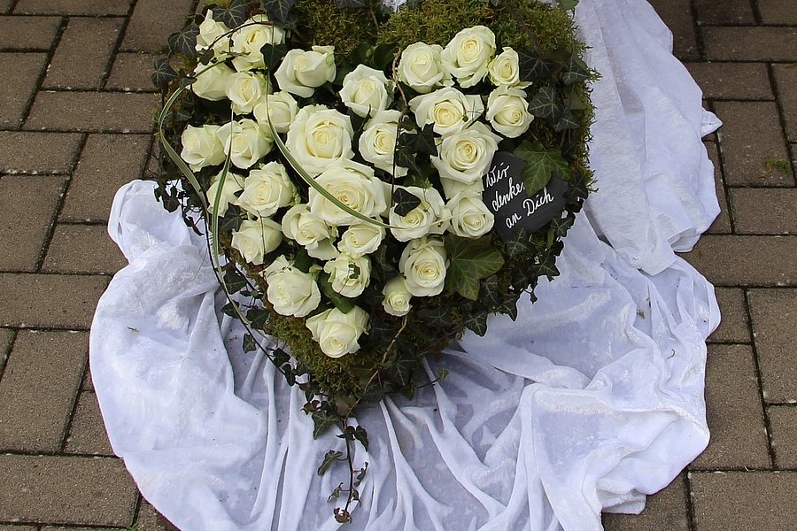Herzförmiger Kranz aus weißen Rosen und Moos, liegend auf weißem Stoff auf gepflastertem Boden.