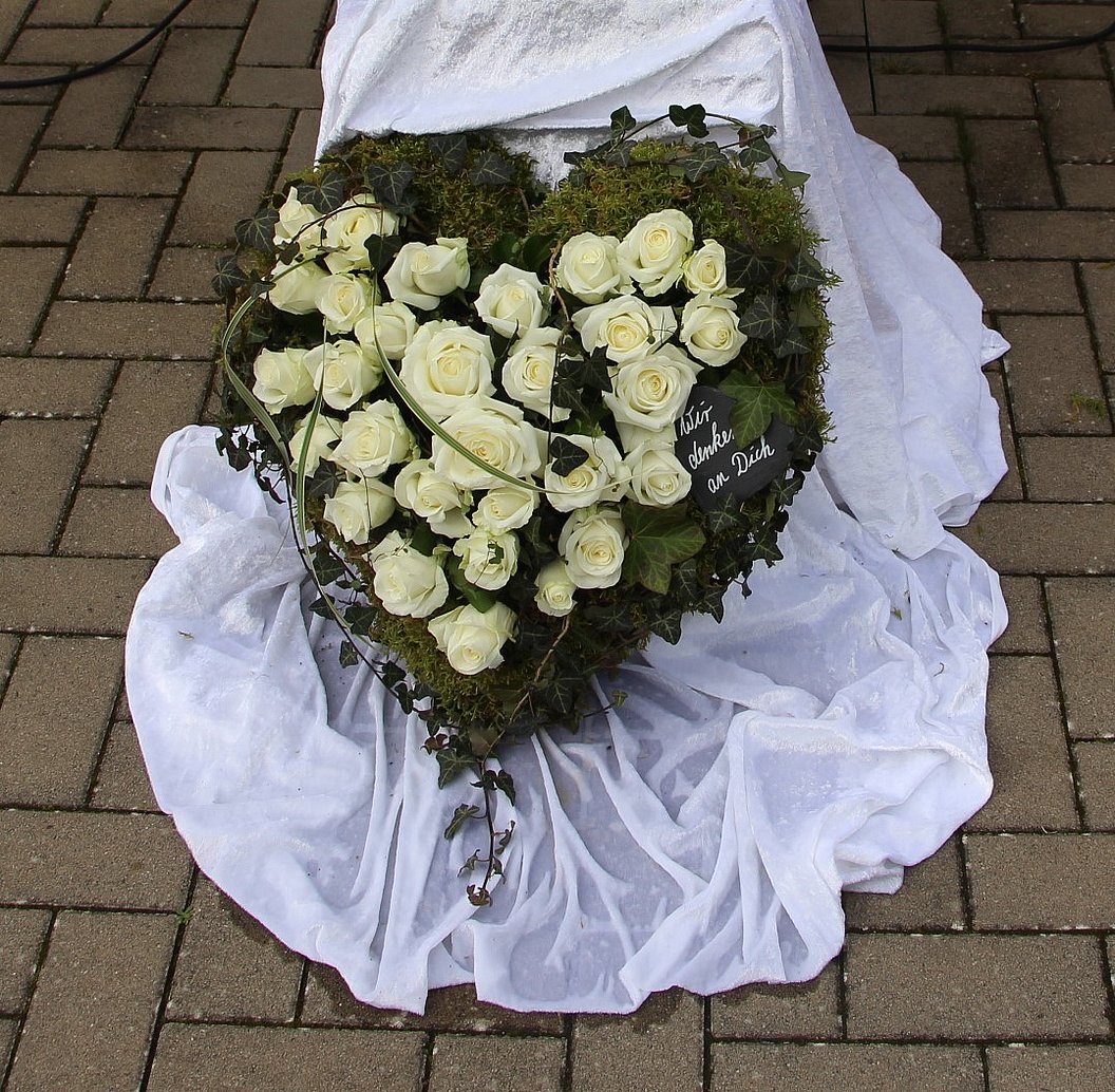 Herzförmiger Kranz aus weißen Rosen und Moos, liegend auf weißem Stoff auf gepflastertem Boden.