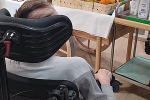 Ansicht eines Rollstuhlfahrers vor einem Verkaufsstand mit Obst und einer Person im Hintergrund.