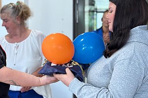Zwei Personen halten gemeinsam ein kleines Modell mit orangefarbenem und blauem Ballon in einem hellen Raum mit Glaswand im Hintergrund.