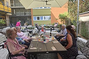 Gruppe älterer Menschen sitzt an einem rechteckigen Gartentisch unter einem großen Sonnenschirm im Innenhof