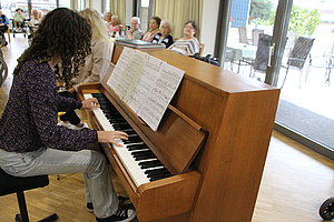 Junge Dame spielt Klavier in einem Raum voller Menschen, Notenblätter sichtbar auf dem Klavier.