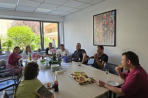Gruppe von acht Personen sitzt an einem Tisch mit Getränken und Snacks in einem hellen Raum mit Fenster und bunter Wandkunst