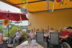 Gruppe älterer Menschen sitzt an mehreren Tischen unter einem gelben Sonnendach mit bunten Dekorationen und hebt teils die Hände