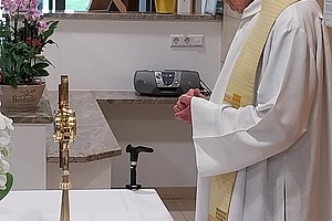 Ein älterer Mann in weißem liturgischem Gewand steht an einem mit weißem Tuch bedeckten Altar in einem Raum mit Fenster und Pflanzen.
