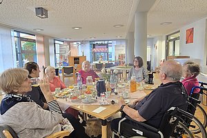 Gruppe von älteren Menschen in einem Café, umgeben von Tischen mit Getränken und Lebensmitteln.