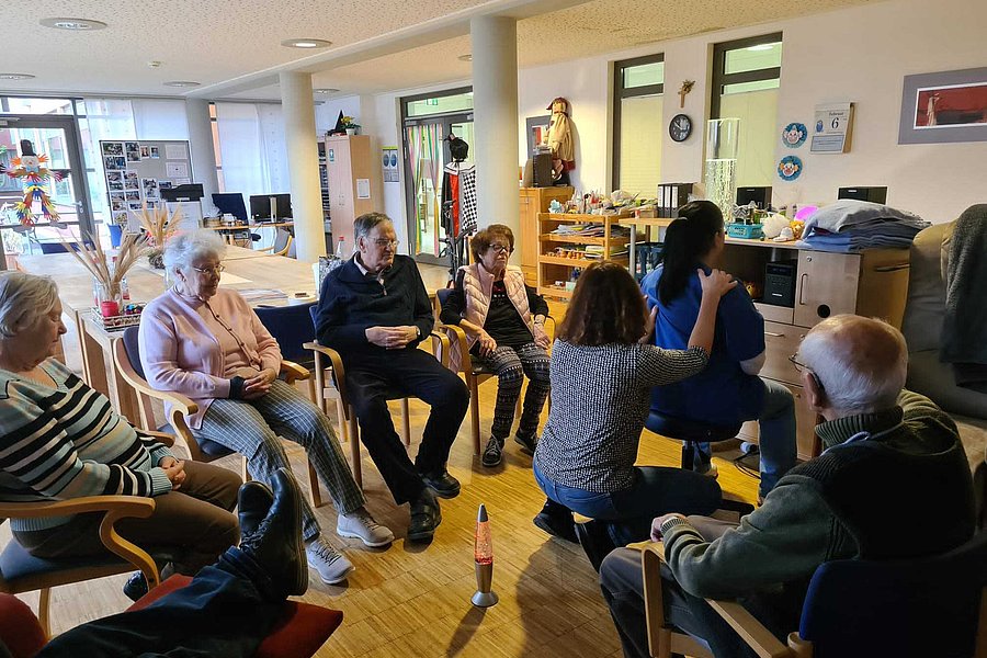 Mehrere ältere Menschen sitzen in einem hellen Raum in einem Stuhlkreis, eine Frau kniet und berührt die Schulter einer sitzenden Frau.