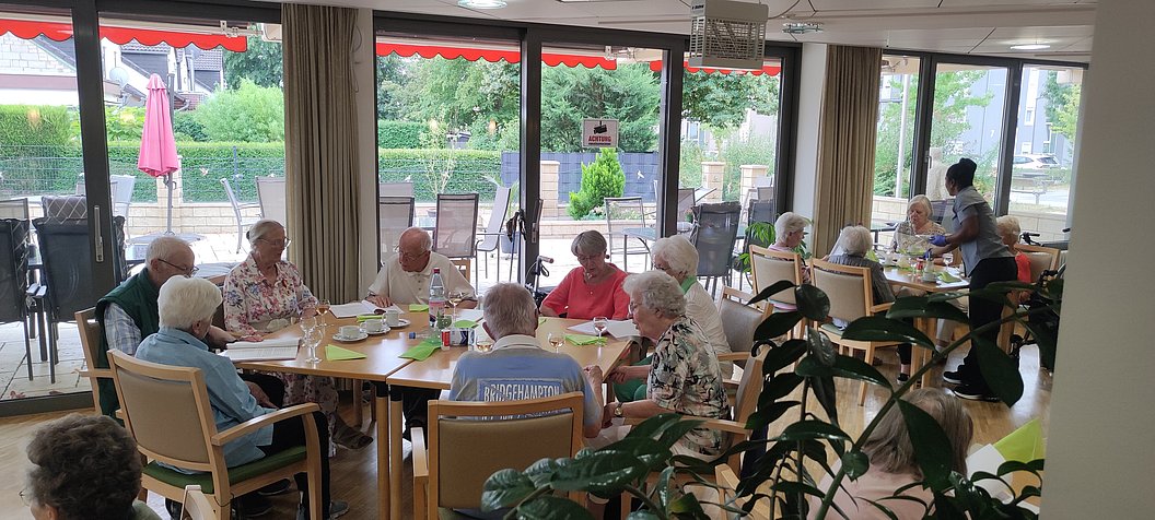 Gruppe älterer Menschen sitzt an einem großen Tisch in einem hellen Raum mit großen Fenstern und Pflanzen