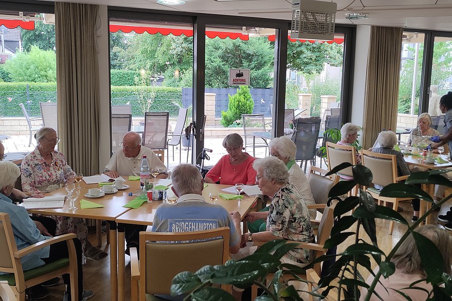 Gruppe älterer Menschen sitzt an einem großen Tisch in einem hellen Raum mit großen Fenstern und Pflanzen