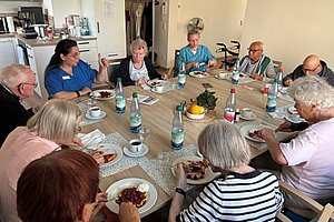Gruppe älterer Menschen sitzt an einem rechteckigen Tisch und isst gemeinsam Mahlzeit mit Wasserflaschen und Kaffeetassen