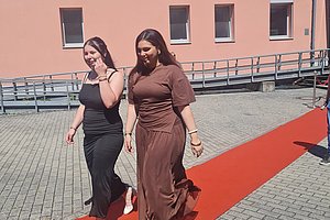 Zwei Frauen gehen nebeneinander auf einem roten Teppich vor einem Gebäude mit oranger Fassade und mehreren Fenstern