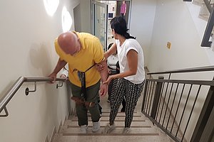 Frau stützt Mann mit Gehstock beim Treppensteigen in einem Treppenhaus