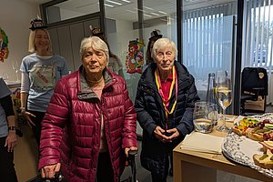 Zwei ältere Frauen mit Winterjacken stehen in einem Raum mit Buffet und Getränken auf einem Tisch