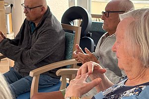 Drei ältere Menschen sitzen nebeneinander auf Stühlen und gestikulieren mit den Händen