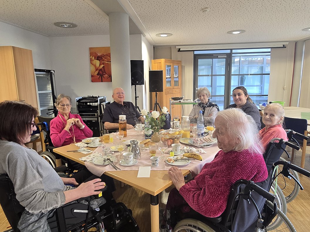 Geselliges Treffen im Aufenthaltsraum mit älteren Menschen, die in Rollstühlen am Tisch sitzen und Getränke genießen.
