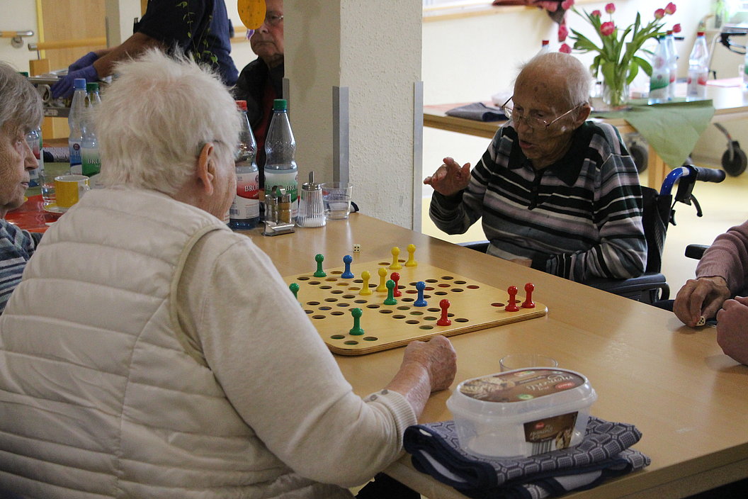 Mehrere ältere Menschen sitzen an einem Tisch und spielen ein Brettspiel mit bunten Spielfiguren.