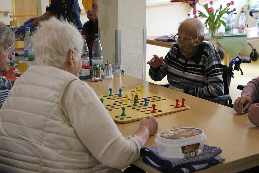 Mehrere ältere Menschen sitzen an einem Tisch und spielen ein Brettspiel mit bunten Spielfiguren.