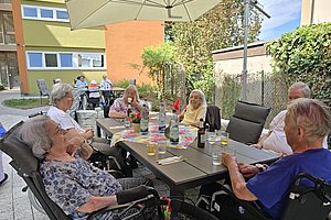 Mehrere ältere Menschen sitzen an einem rechteckigen Gartentisch im Freien unter einem großen Sonnenschirm und unterhalten sich
