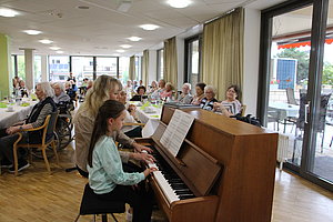 Musikaufführung am Klavier mit einem Kind und einer Frau in einem Saal voller Senioren.