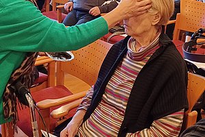 Szenenbild einer Seniorengruppe, eine Betreuerin hilft einer älteren Dame in einem Aufenthaltsraum.
