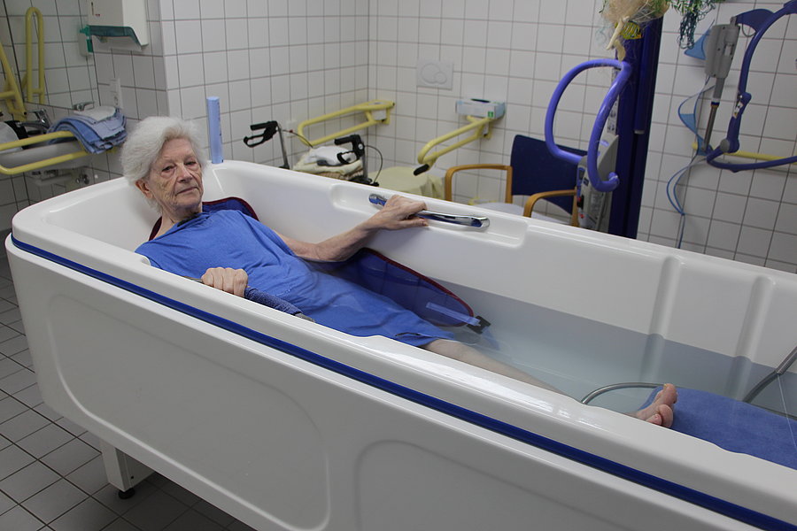 Ältere Person liegt in einer weißen Badewanne mit blauer Umrandung in einem gefliesten Raum und hält sich am Badewannenrand fest