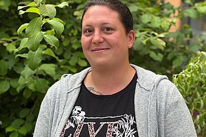 Frau mit zusammengebundenem Haar trägt grauen Kapuzenpullover über schwarzem T-Shirt mit floralem Schriftzug und steht vor grünem Laub und einem Gebäude.