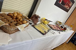 Buffet mit Körben voller Brötchen und Croissants, Tabletts mit Käse, Aufschnitt und Schalen mit Butter und Marmelade auf weiß gedecktem Tisch