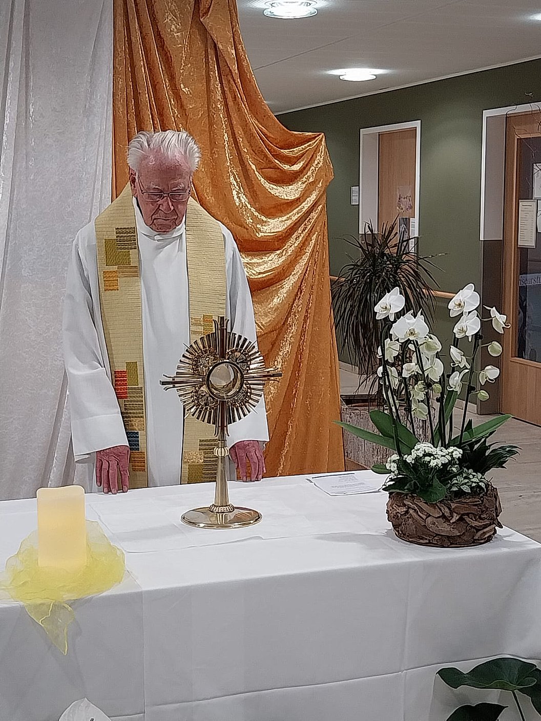 Ein Priester in weißem Gewand und Stola steht hinter einem mit weißem Tuch bedeckten Altar mit einem goldenen Monstranz-Objekt und einem Blumenarrangement aus weißen Orchideen