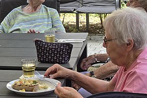 Ältere Frau mit Brille sitzt im Rollstuhl an einem Gartentisch und isst von einem Teller mit Besteck